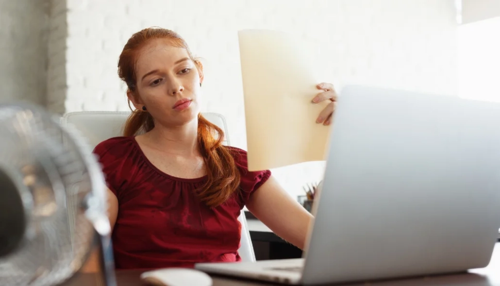 Young lady at a desk with a fan in front of her, fanning herself with a piece of paper